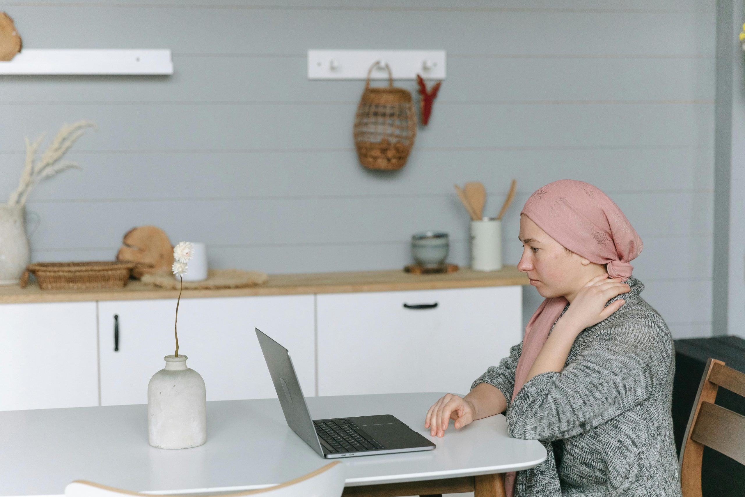 Woman with head wrap sitting at a kitchen table, using a laptop, symbolizing resilience.
